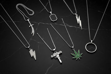 Flat lay arrangement of eight silver pendant chains from The Dazzle, featuring minimalist and unique charms like the Zulfiqar sword, Uzi gun, lightning bolt, ring, geometric bar, and cannabis leaf pendants, styled on a clean background.