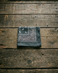 Slate gray square bandana with white paisley pattern folded on a dark weathered wood surface – Dazzle classic collection