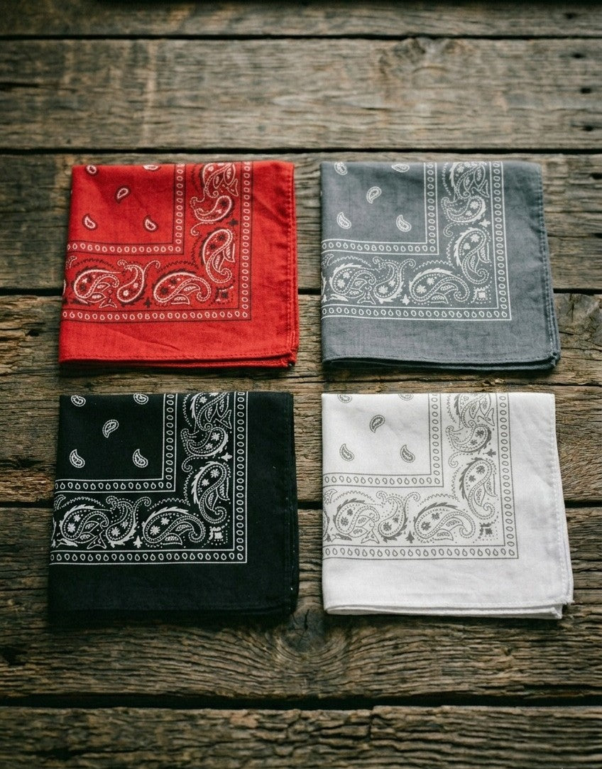 Flat lay of four folded Dazzle paisley bandanas in red, gray, black, and white arranged together on a rustic wooden surface