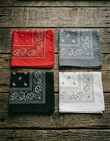 Flat lay of four folded Dazzle paisley bandanas in red, gray, black, and white arranged together on a rustic wooden surface