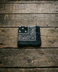 Midnight black premium cotton bandana with white paisley details folded on an organic wood plank background – Dazzle fashion accessories.